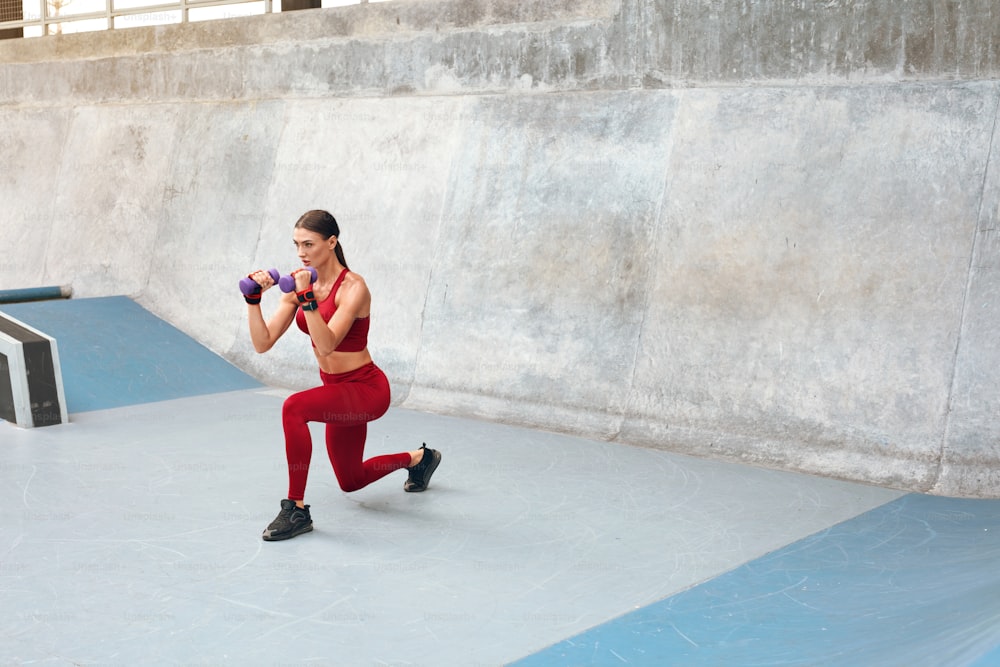 Deporte. Mujer se pone en cuclillas contra la pared de concreto. Chica fitness con cuerpo musculoso fuerte en traje deportivo de moda haciendo ejercicio en el estadio al aire libre. Estilo de vida urbano y personas activas.
