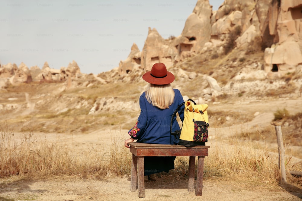 Travel. Woman Traveler In Hat With Backpack Traveling To Cave Town, Sitting On Bench Near Rocks. High Resolution