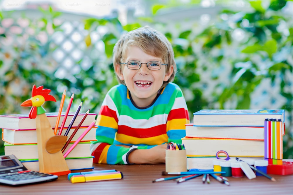 Cute beautiful little kid boy with glasses and lots of pupil's stuff like crayons pens, scissors and books. Child and student is back to school and happy about begin of lessons.