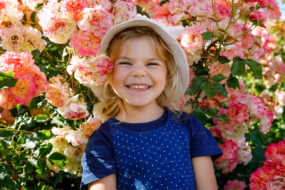 Porträt eines kleinen Kleinkindes im blühenden Rosengarten. Nettes schönes schönes Kind, das Spaß mit Rosen und Blumen in einem Park an einem sonnigen Sommertag hat. Glückliches lächelndes Baby