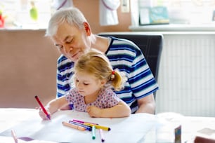 Cute little baby toddler girl and handsome senior grandfather painting with colorful pencils at home. Grandchild and man having fun together. Family and generation in love.