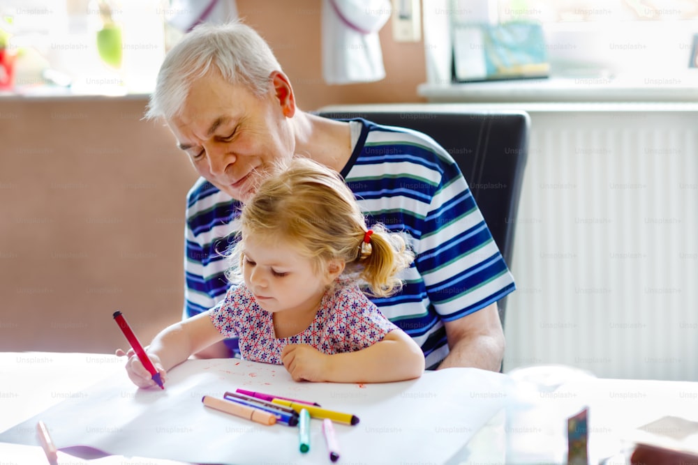 Cute little baby toddler girl and handsome senior grandfather painting with colorful pencils at home. Grandchild and man having fun together. Family and generation in love.