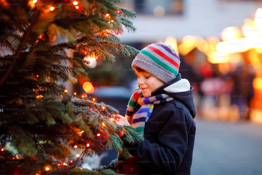 Pequeño niño lindo divirtiéndose en el mercado tradicional de Navidad durante fuertes nevadas. Niño feliz disfrutando del mercado familiar tradicional en Alemania. Colegial de pie junto al árbol de Navidad iluminado