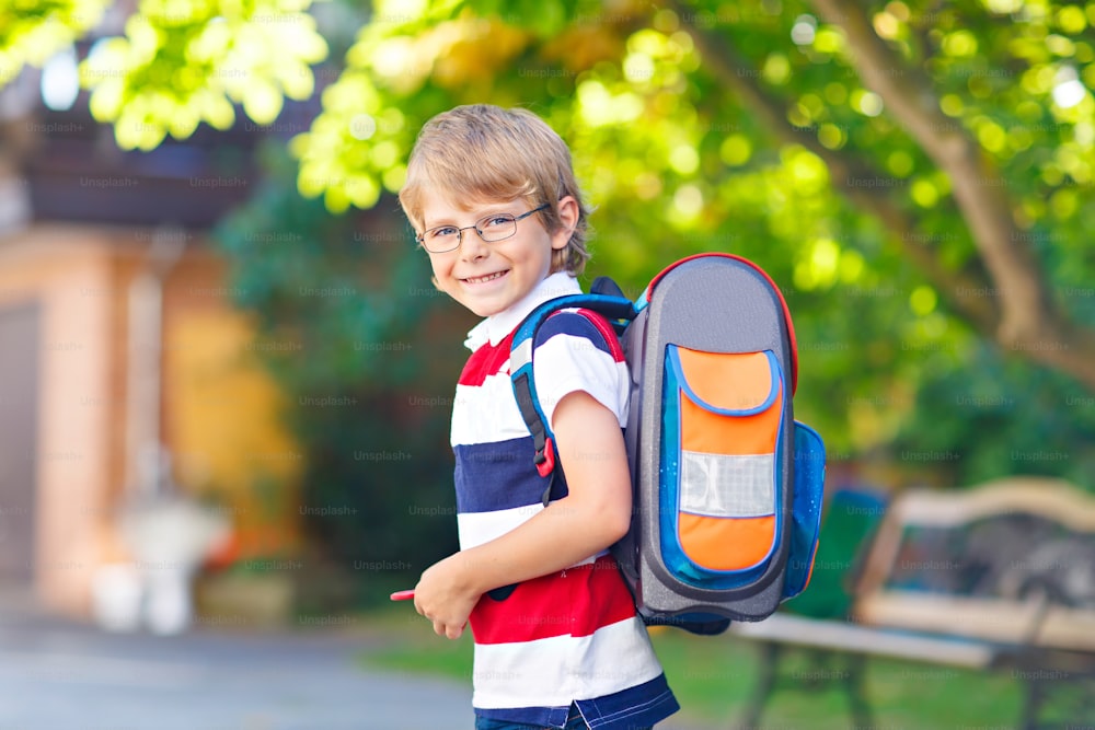Happy little kid boy with glasses and backpack or satchel on his first day to school or nursery. Child outdoors on warm sunny day, Back to school concept