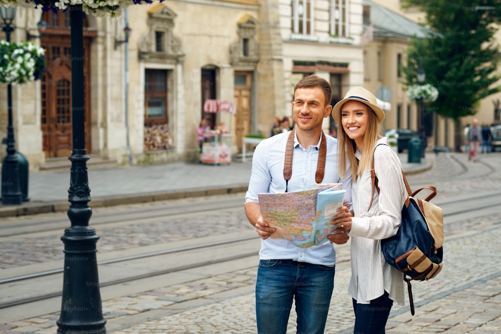 Tourist Couple With Map Walking On City Street. Young Smiling People Traveling On Weekend Vacation, Walking Around Old Town Streets And Looking At Architecture. Travel Concept. High Quality Image.