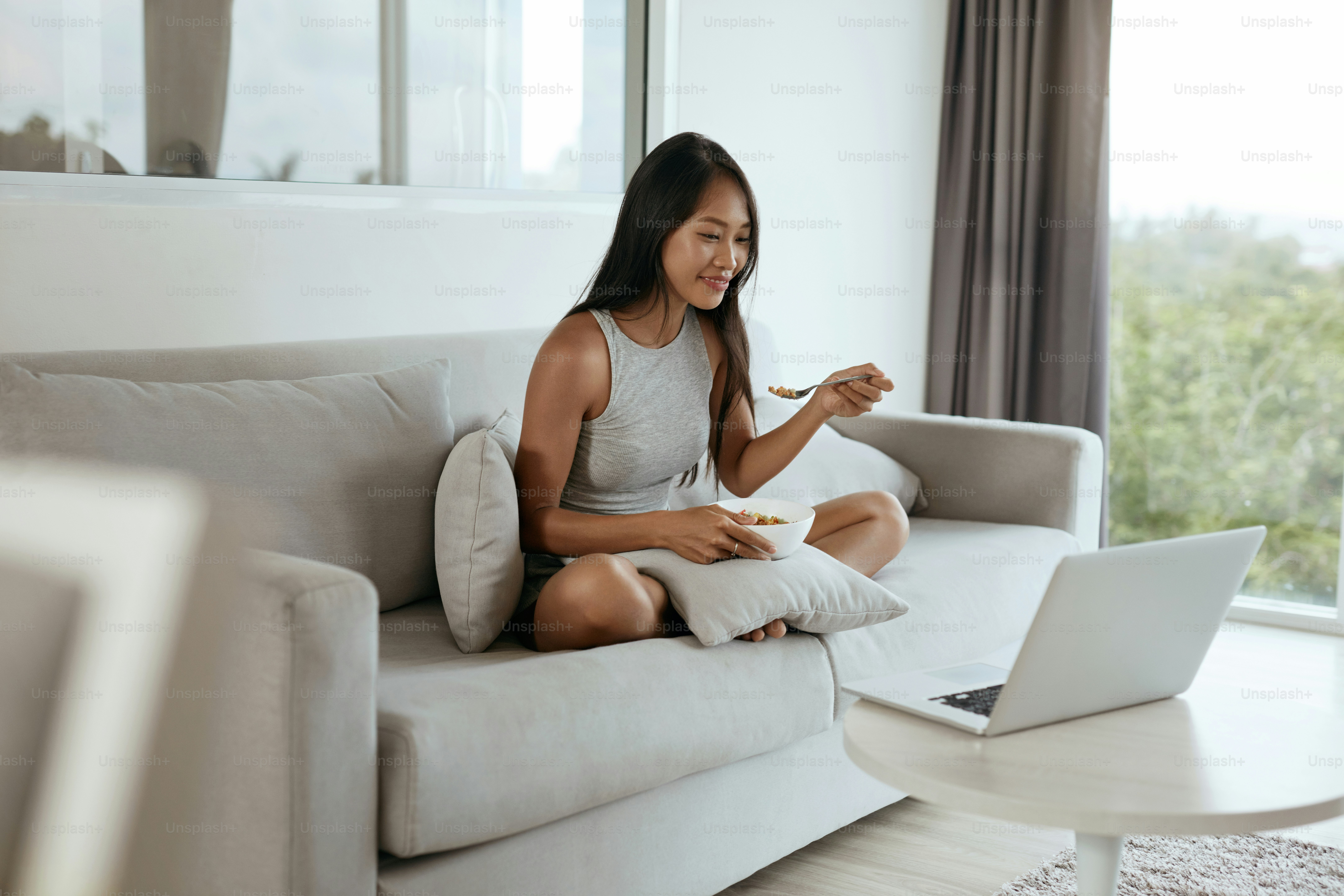 Mujer desayunando mientras usa la computadora en casa por la mañana. Hermosa chica asiática sonriente comiendo comida relajándose en el sofá con computadora portátil en la sala de estar luminosa