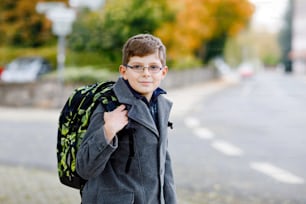 Glücklicher kleiner Junge mit Brille und Rucksack oder Schulranzen. Schulkind in stilvollem Fashon-Coan auf dem Weg zur Mittel- oder Oberschule an einem kalten Herbsttag. Gesundes Kind draußen auf der Straße, an regnerischen Tagen