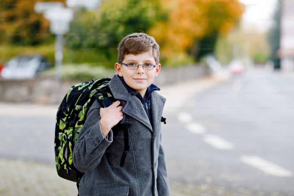 Happy kid boy with glasses and backpack or satchel. Schoolkid in stylish fashon coan on the way to middle or high school on cold autumn day. Healthy child outdoors on the street, on rainy day