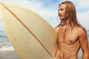 서퍼 초상화. 하얀 서핑보드를 들고 있는 긴 젖은 머리를 가진 잘생긴 서핑 남자. 활동적인 라이프스타일을 위한 수상 스포츠.