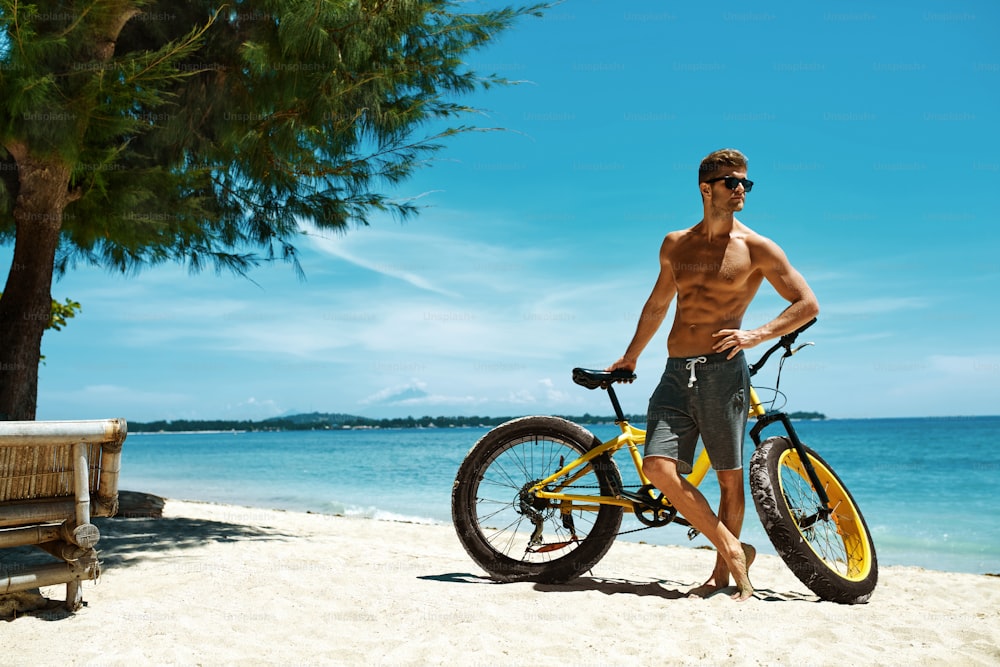 夏の旅行ビーチでの休暇で海岸でリラックスする黄色い砂の自転車を持つハンサムな筋肉質の男性。海で日光浴をする自転車を持つフィットネス男性モデル。夏のコンセプトにおけるスポーツ活動とレクリエーション