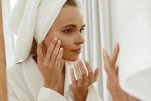 Chica europea aplicando un parche debajo del ojo en la cara y mirar en el espejo. Joven mujer hermosa concentrada usa bata de baño y toalla de baño envuelta en la cabeza. Concepto de cuidado de la piel facial