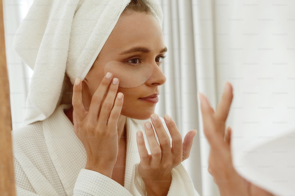 European girl applying under eye patch on face and watch in mirror. Young concentrated beautiful woman wear bathrobe and wrapped bath towel on head. Concept of face skin care