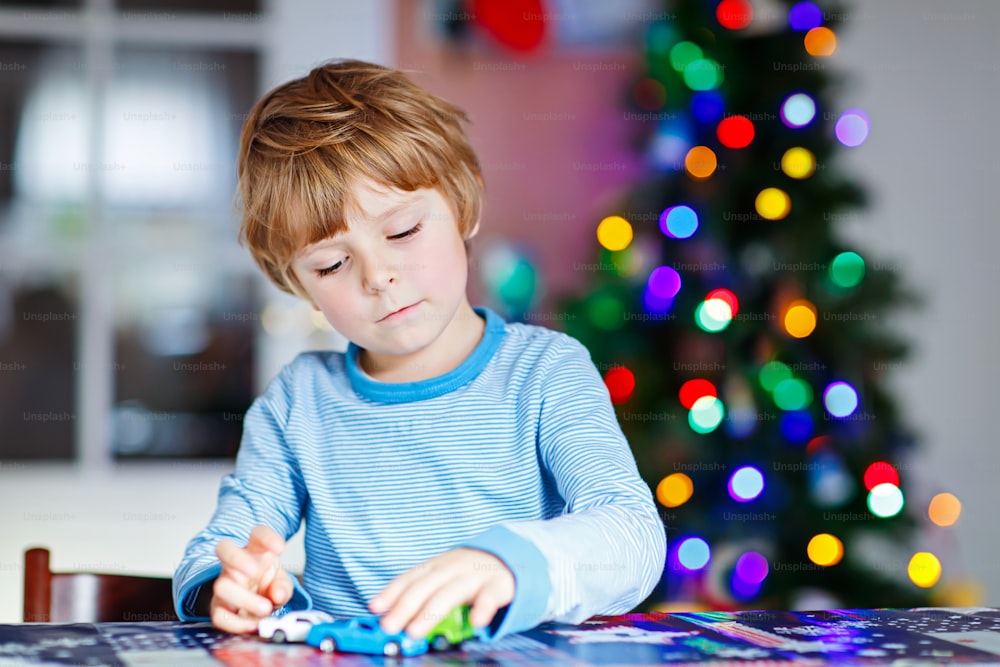 Little blond child playing with cars and toys at home, indoor. Cute happy funny kid boy having fun with gifts. Colorful lights on background. Christmas time concept.