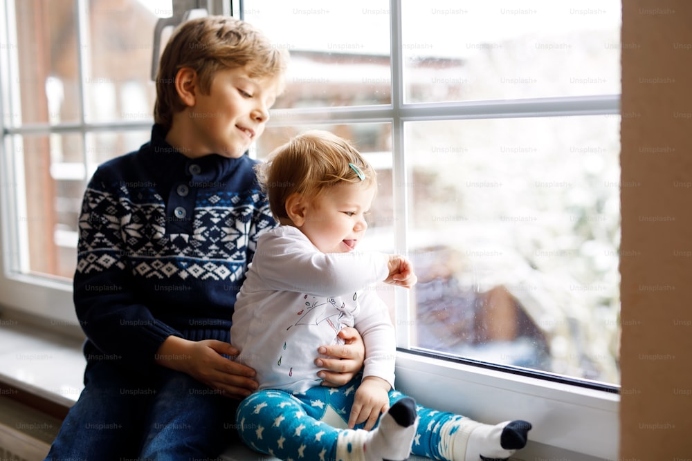 행복한 사랑스러운 소년과 귀여운 아기 소녀가 창가에 앉아 크리스마스 날이나 아침에 눈 위를 바라보고 있습니다. 웃고 있는 아이들, 형제자매, 여동생, 남동생들이 겨울 눈을 바라보고 있다