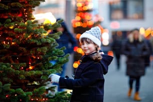 Little cute kid girl having fun on traditional Christmas market during strong snowfall. Happy child enjoying traditional family market in Germany. Schoolgirl standing by illuminated xmas tree