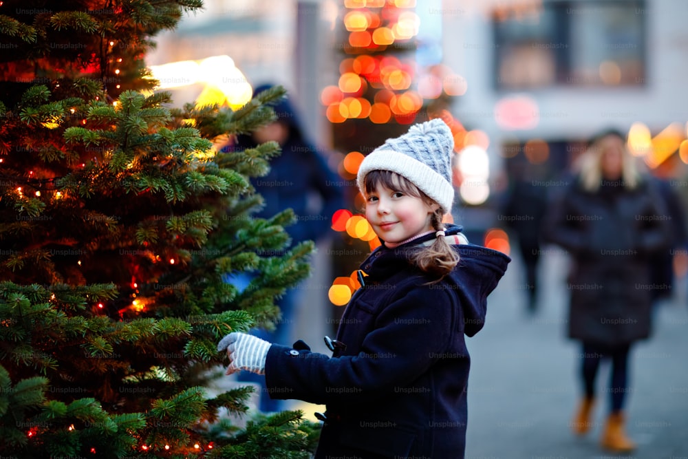 Menina criança bonito se divertindo no mercado tradicional de Natal durante a forte queda de neve. Criança feliz desfrutando do mercado familiar tradicional na Alemanha. Estudante em pé ao lado da árvore de Natal iluminada