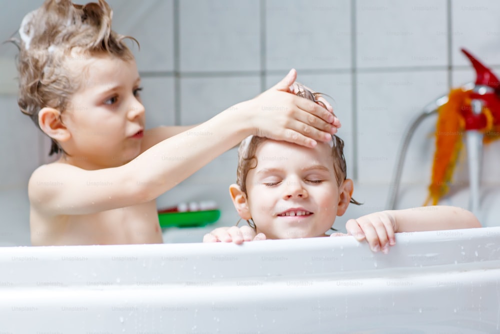 Happy siblings: Two little twins children playing together with water by taking bath in bathtub at home. Kid boys having fun together, helping with hair washing.