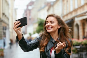 Videollamadas de mujer por teléfono en la calle. Mujer joven sonriente tomando fotos en el teléfono móvil al aire libre. Alta resolución