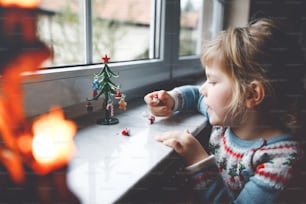 Niña pequeña sentada junto a la ventana y decorando un pequeño árbol de Navidad de vidrio con pequeños juguetes navideños. Feliz niño sano celebra la fiesta tradicional familiar. Bebé adorable