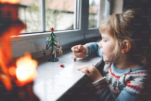 Litte toddler girl sitting by window and decorating small glass Christmas tree with tiny xmas toys. Happy healthy child celebrate family traditional holiday. Adorable baby