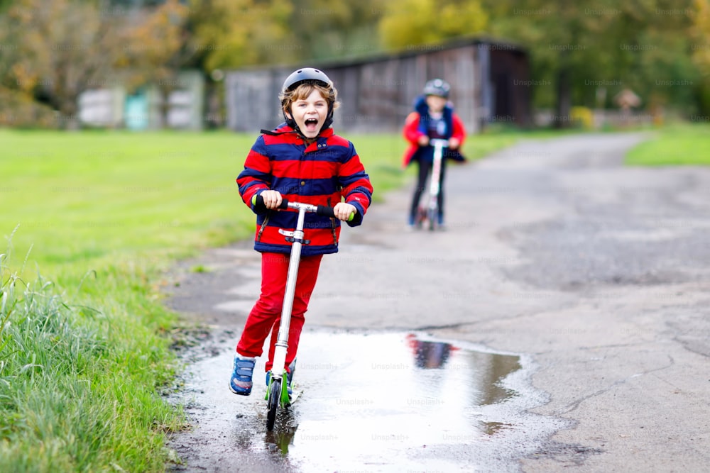 Due bambini piccoli che cavalcano su scooter a spinta sulla strada da o verso la scuola. Scolari di 7 anni che guidano attraverso la pozzanghera di pioggia. Fratelli divertenti e migliori amici che giocano insieme
