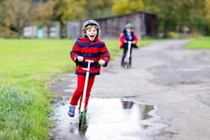 Two little kids boys riding on push scooters on the way to or from school. Schoolboys of 7 years driving through rain puddle. Funny siblings and best friends playing together