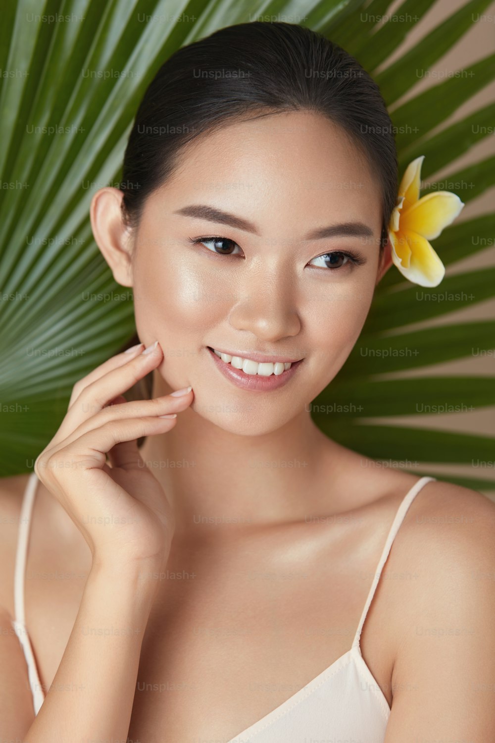 Belleza. Retrato de fondo de mujer sobre hoja. Hermosa modelo asiática con flor en el cabello tocando la cara y mirando hacia otro lado. Mujer joven con piel perfecta y maquillaje natural después de usar cosméticos orgánicos.