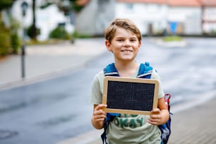 Happy little kid boy with backpack or satchel. Schoolkid on the way to middle or high school. Child outdoors on the street. Back to school. Kid holding empty chalk desk.