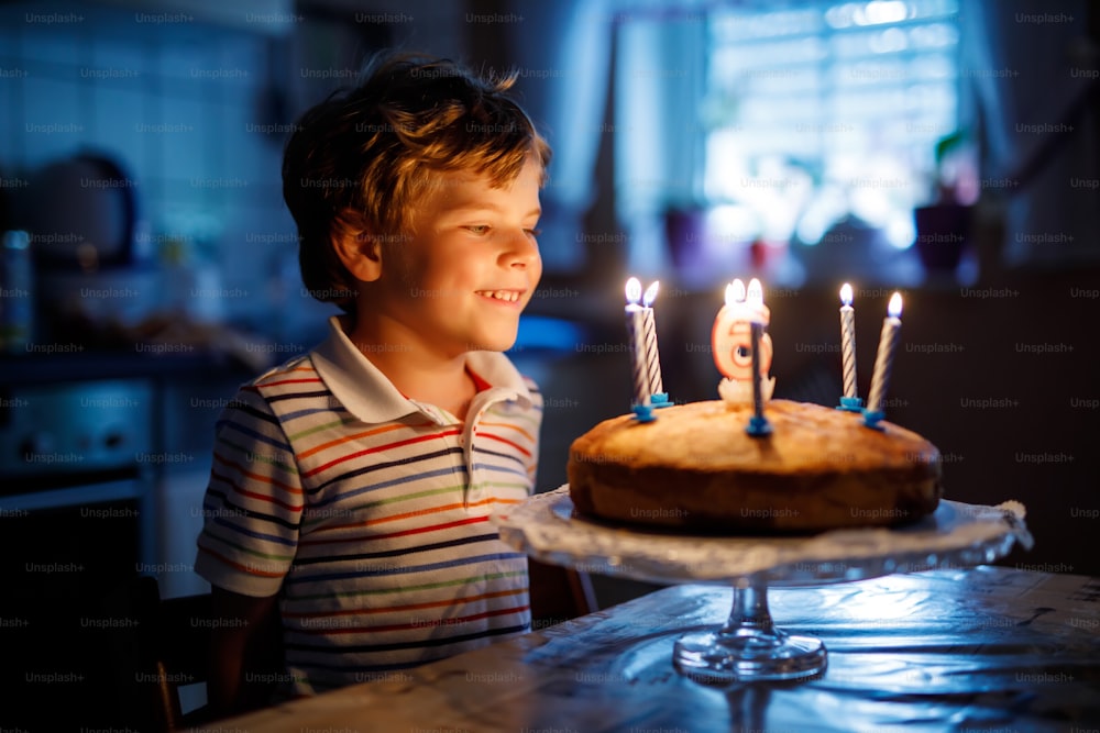 Adorable happy blond little kid boy celebrating his birthday. Child blowing candles on homemade baked cake, indoor. Birthday party for school children, family celebration of 6 years.