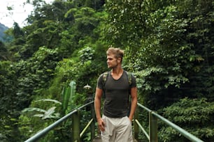 バックパックを持つ観光客が橋の上に立ち、緑のジャングルの森で自然を楽しむ。夏休みに旅行するハンサムな若い男性旅行者。旅行と観光のコンセプト。