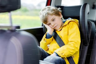 交通渋滞中に車に座っている疲れた就学前の少年。ベルト付きの安全運転席に座った悲しい小さな小学生で、旅行とジョニーを楽しんでいます。子供との安全な旅行と交通法のコンセプト。