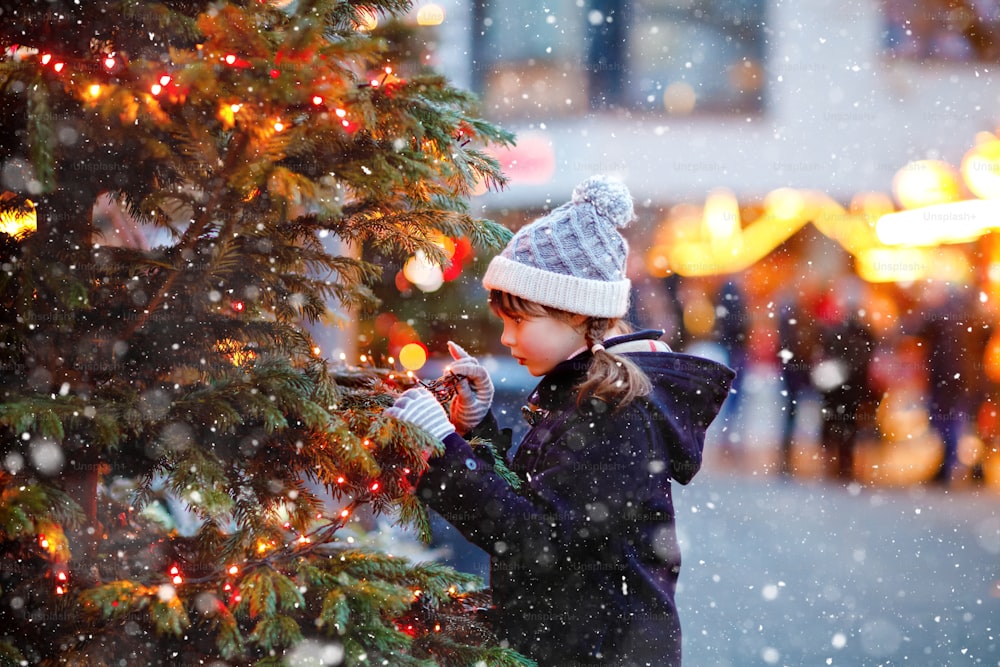 Little cute kid girl having fun on traditional Christmas market during strong snowfall. Happy child enjoying traditional family market in Germany. Schoolgirl standing by illuminated xmas tree