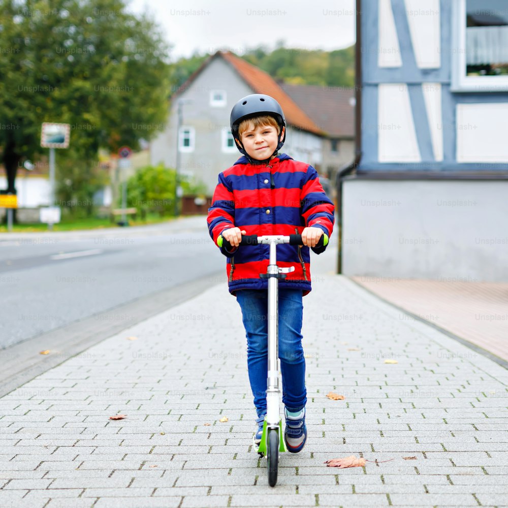 Lindo niño de la escuela con casco montado en scooter en la naturaleza del parque. Actividades infantiles al aire libre en invierno, primavera u otoño. Niño feliz divertido con ropa de moda colorida
