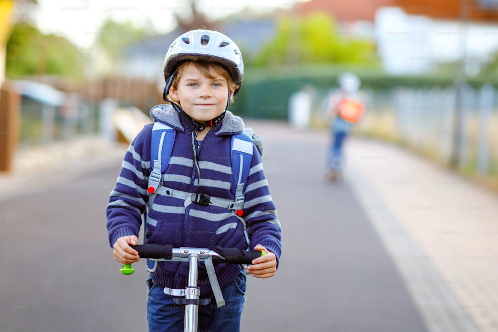 Garoto ativo da escola em capacete de segurança andando com sua scooter na cidade com mochila em dia ensolarado. Criança feliz em roupas coloridas pedalando a caminho da escola. Maneira segura para as crianças ao ar livre para a escola.