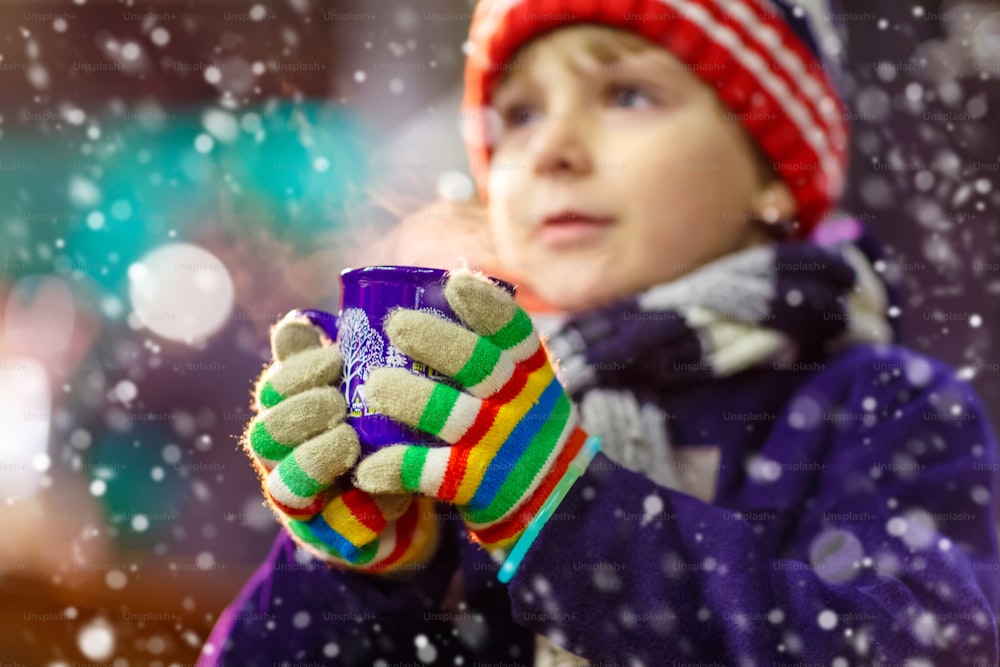 Kleiner süßer Junge mit einer Tasse dampfender heißer Schokolade oder Kinderpunsch. Glückliches Kind auf dem Weihnachtsmarkt in Deutschland. Traditionelle Freizeit für Familien an Weihnachten