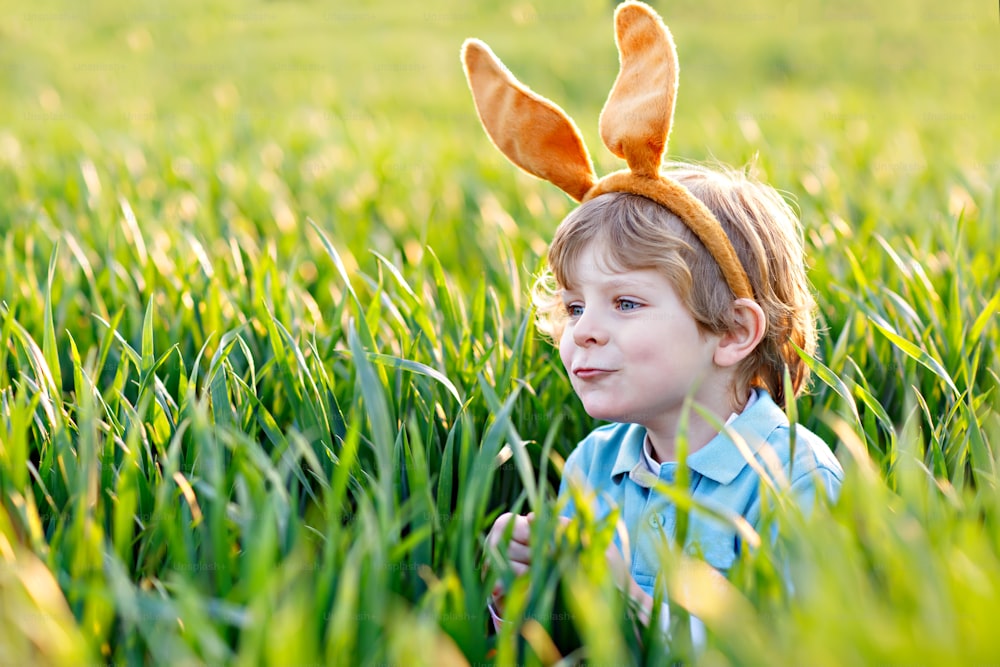 Mignon petit garçon avec des oreilles de lapin s’amusant avec la chasse traditionnelle aux œufs de Pâques par une chaude journée ensoleillée, à l’extérieur. Célébrer les vacances de Pâques. Découverte de tout-petits, œufs colorés dans l’herbe verte.
