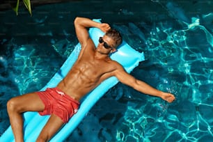Homme Mode d’été. Bel homme avec un corps sexy en maillot de bain, des lunettes de soleil à la mode bronzant, flottant dans l’eau de la piscine au Relax Spa Resort. Modèle de fitness avec bronzage de la peau relaxant en vacances