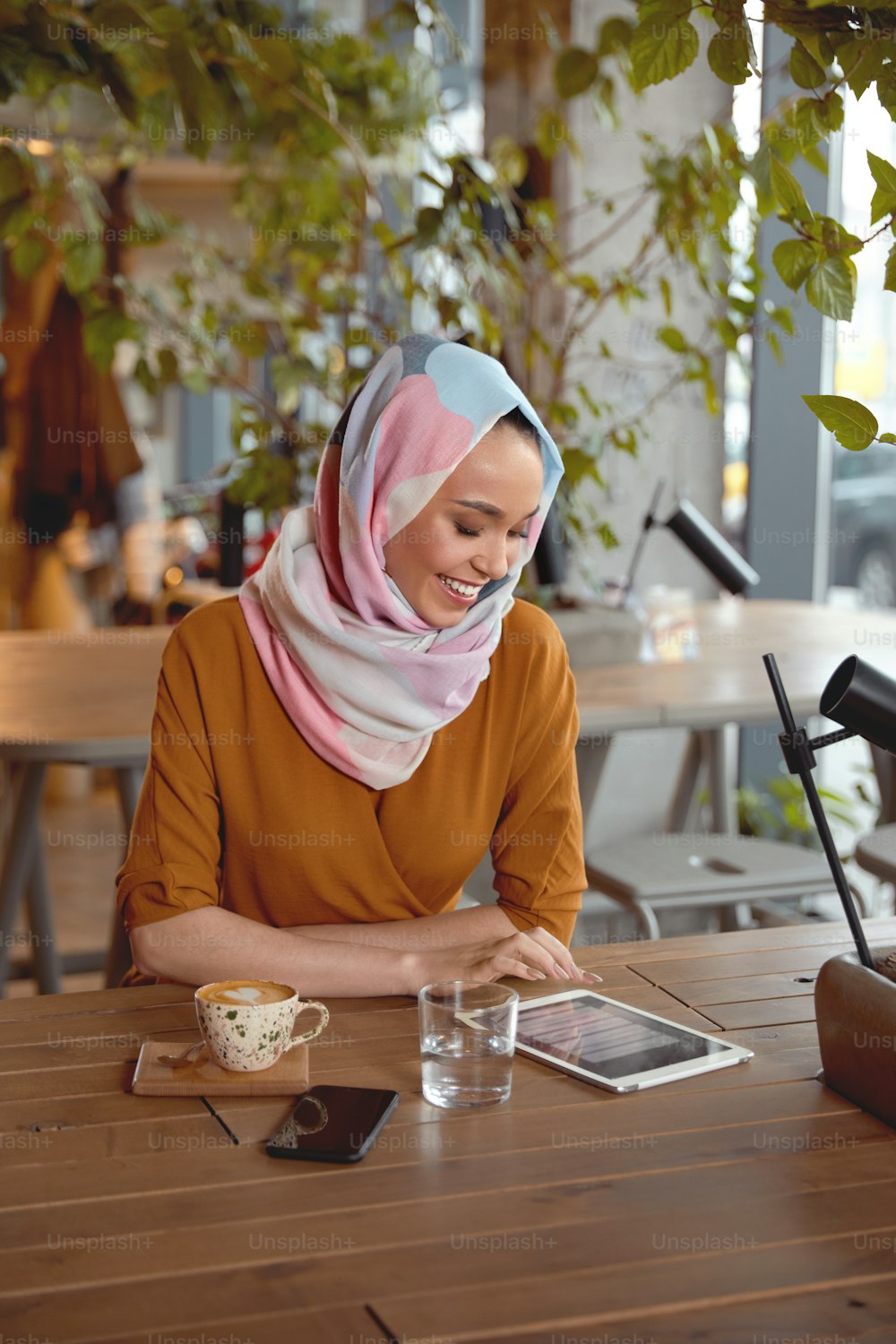 ヒジャーブの女の子。若いイスラム教徒の女性のポートレート。カフェに座ってタブレットで作業する美しい女性。画面を見て読む笑顔のモデル。