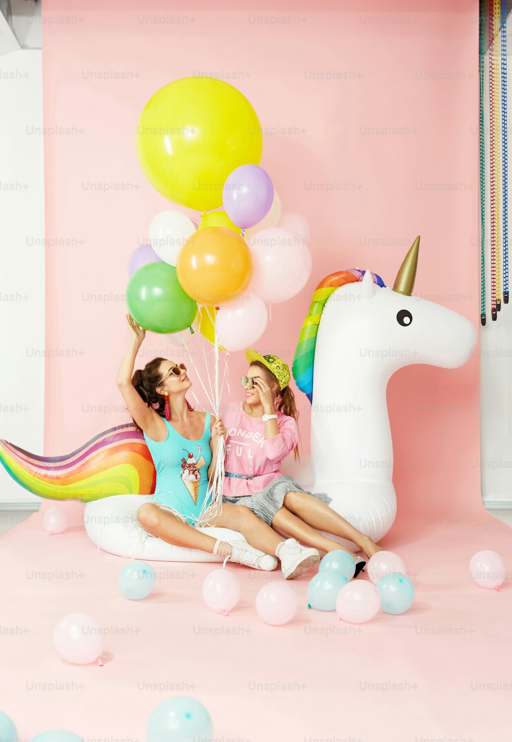 Chicas de la moda de verano divirtiéndose con globos en Unicorn Float. Hermosas mujeres sonrientes con ropa de moda y trajes de baño con globos de colores sobre fondo rosa. Estilo de mujeres. Imagen de alta calidad.