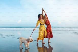 Verano. Mujeres con perro en la playa. Fashion Girls en maxi boho vestidos caminando descalzos con mascota a lo largo del océano tropical. Modelos felices en ropa de moda en la costa que admite perros en vacaciones de verano.