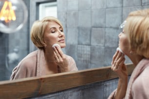 Rotina de Skincare. Conceito de Tratamento de Beleza Anti Envelhecimento. Mulher madura limpa a pele do rosto com almofadas de algodão na frente do reflexo do espelho.