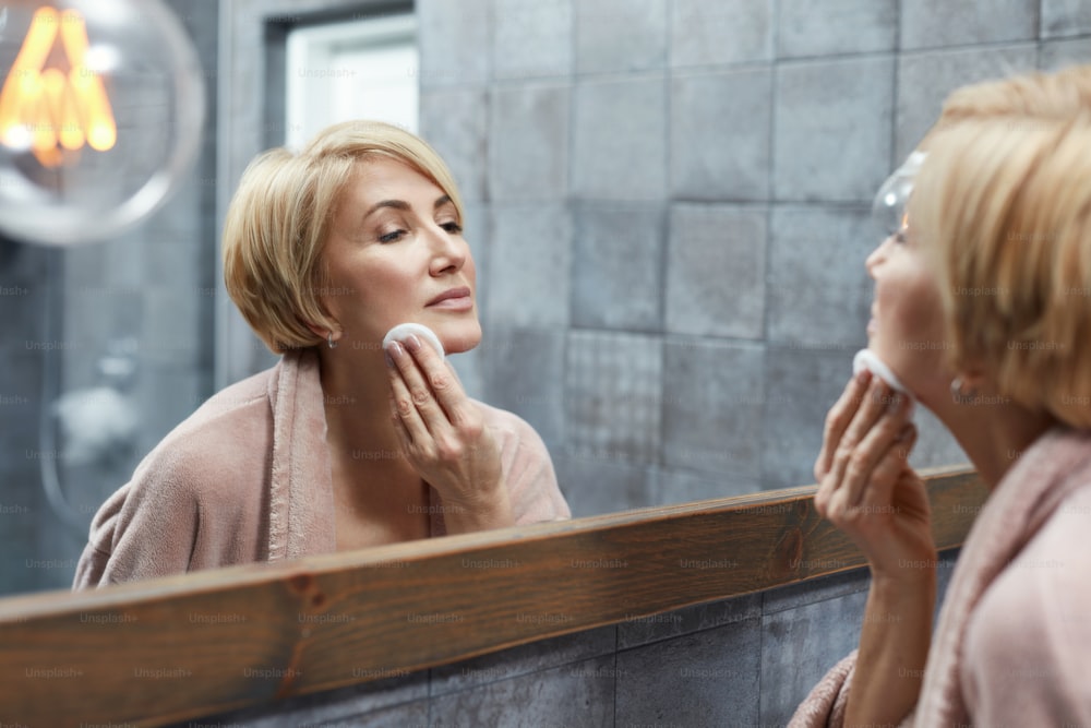 Skincare Routine. Anti Aging Beauty Treatment Concept. Mature Woman Cleans Face Skin With Cotton Pads In Front Of The Mirror Reflection.