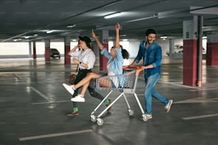 Young People Having Fun, Racing On Shopping Trolley At Parking, Happy Friends With Cart. High Resolution