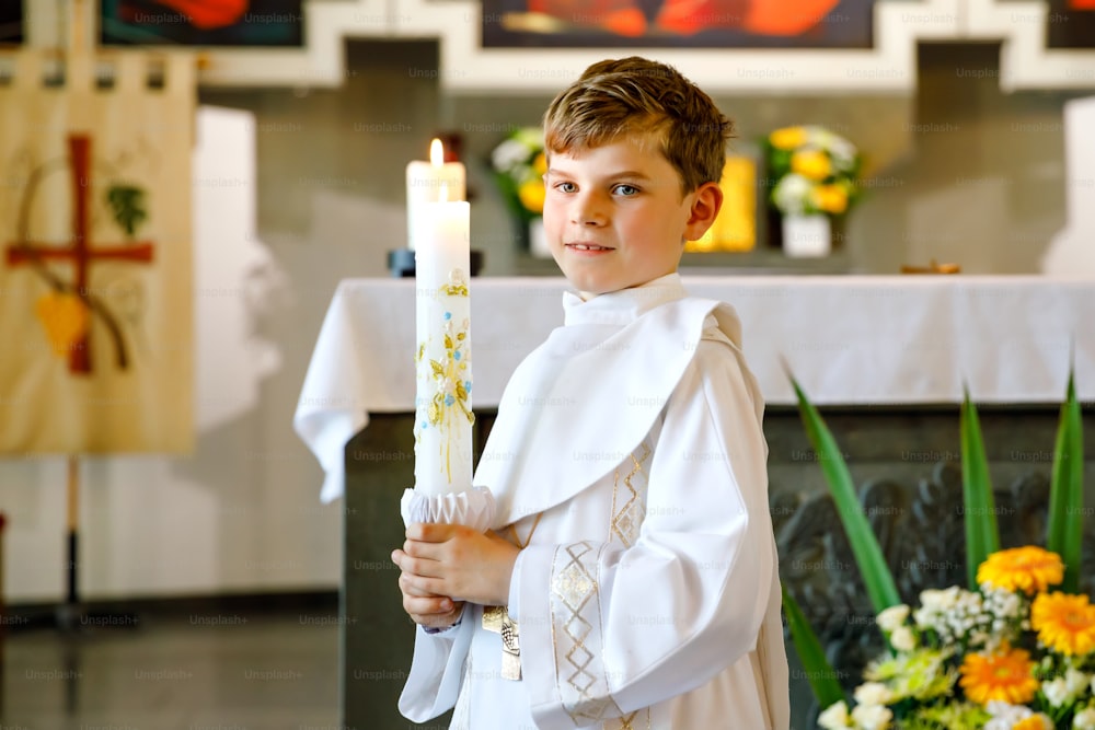 첫 번째 성찬식을 받는 어린 소년. 세례 촛불을 들고 있는 행복한 아이. 가톨릭 교회의 전통. 제단 근처 교회에서 하얀 전통 가운을 입은 아이