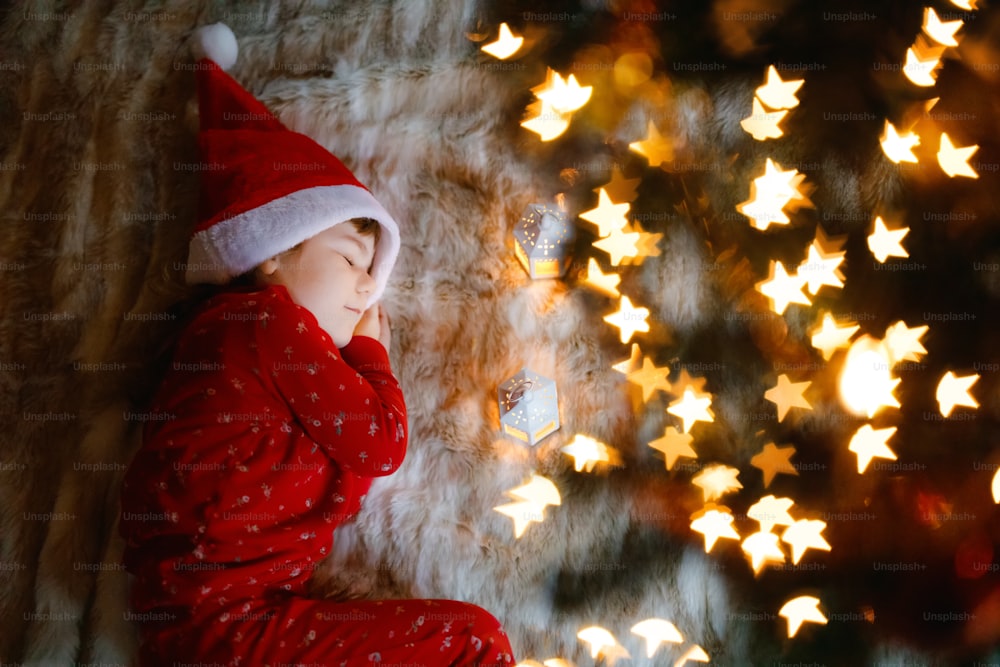 Little cute toddler girl in pajamas under Christmas tree and dreaming of Santa at home, indoors. Traditional Christian family festival. Happy preschool child waiting for gifts on xmas. Cozy soft light.