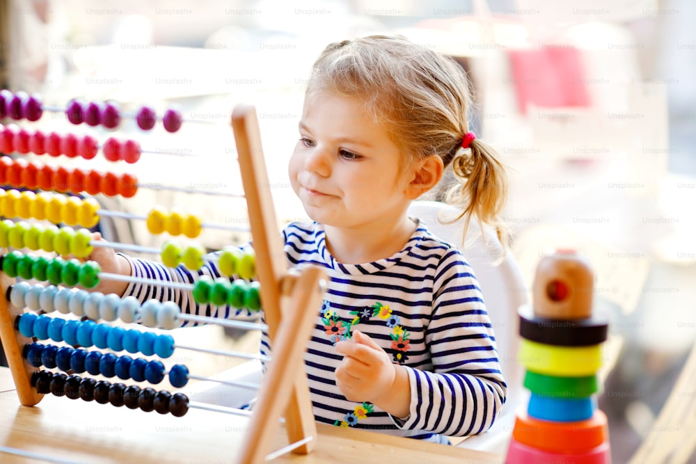 教育的な木製の虹のおもちゃのピラミッドとカウンターそろばんで遊ぶ愛らしいかわいい美しい幼児の女の子。晴れた日に屋内で数えて色を学ぶ健康な幸せな赤ちゃん