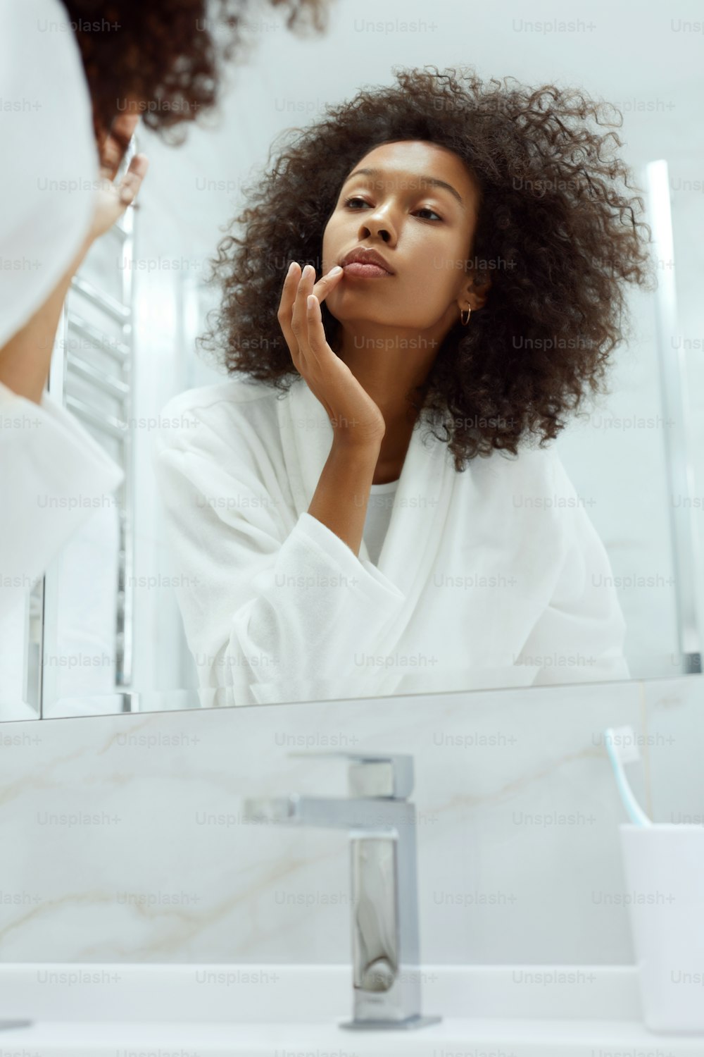 Lips skin care. Woman applying lip balm looking in mirror at bathroom. Portrait of beautiful african girl model with beauty face and natural makeup applying lip product with finger