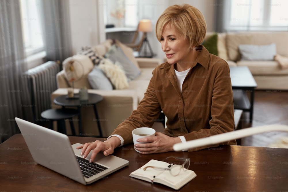 Mujer con computadora portátil. Concepto de tarea remota. Mujer madura con chaqueta marrón lee de la computadora y bebe de la taza.
