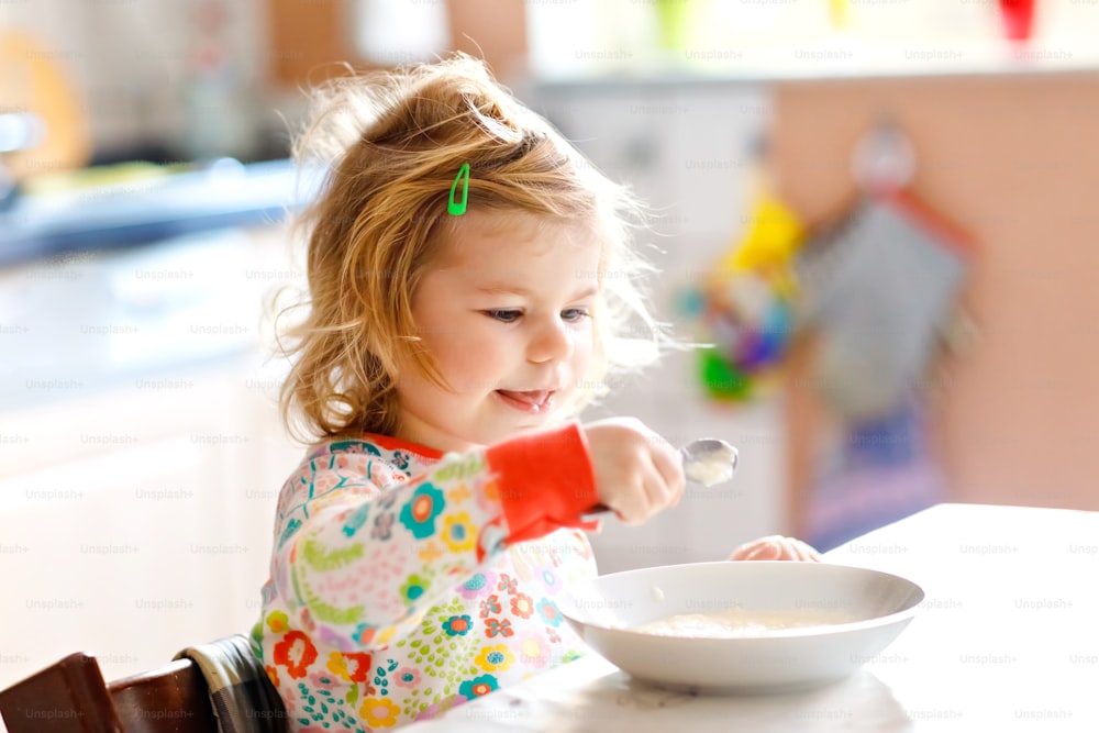 Adorable toddler girl eating healthy porrige from spoon for breakfast. Cute happy baby child in colorful pajamas sitting in kitchen and learning using spoon