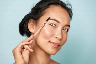 Beauty. Smiling woman brushing eyebrows with makeup brush closeup. Portrait of beautiful happy asian girl model doing make up, shaping brows with cosmetic brush in studio.
